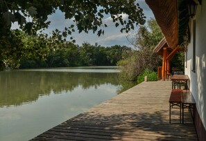 A wooden deck with benches overlooking a calm lake, surrounded by trees and a building with a thatched roof. - Antal-tavi Fogadó és Horgászparadicsom (Szentes)
