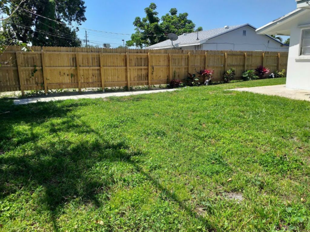 A backyard with a wooden fence, a lawn, and a house in the background.