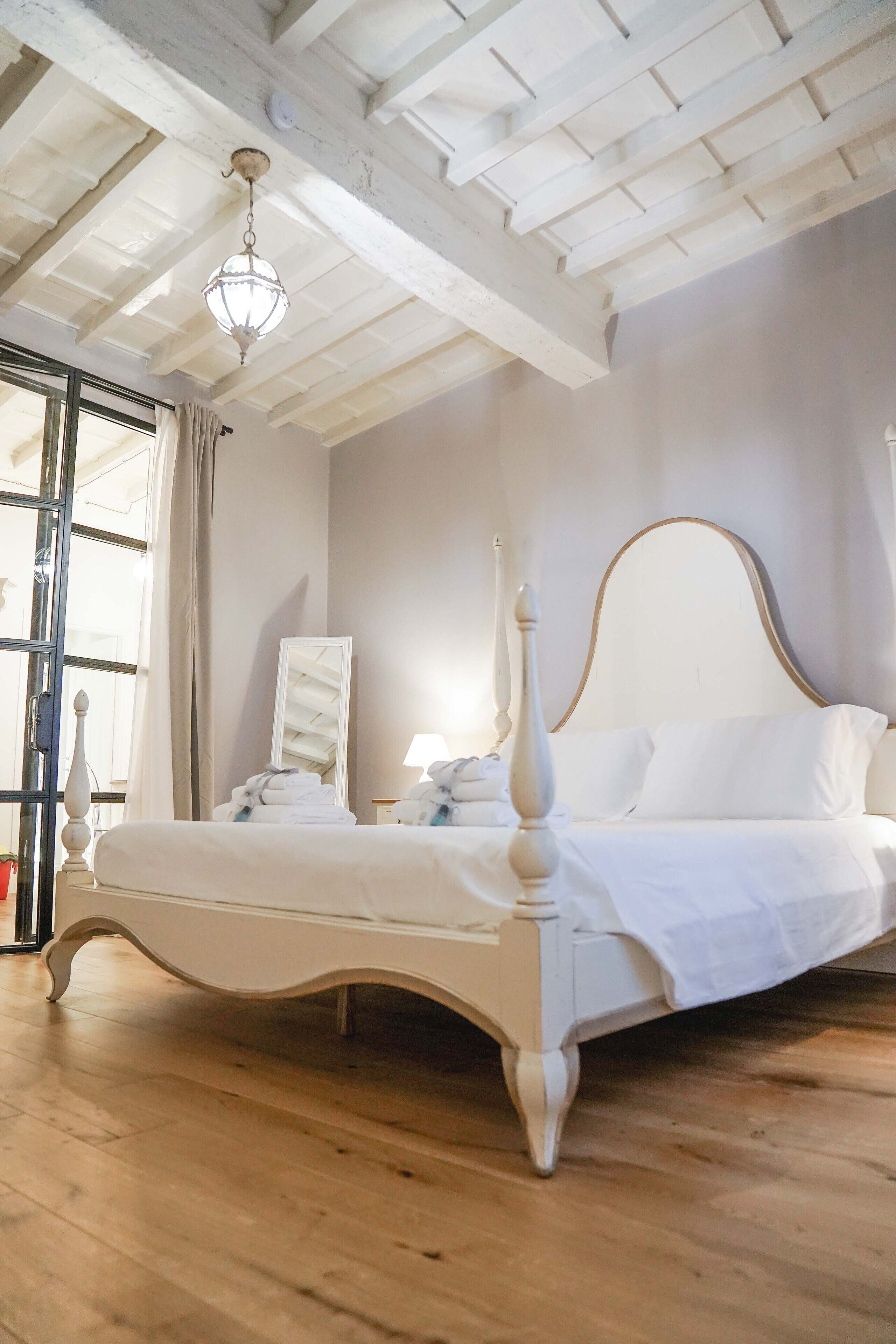 A white four-poster bed with a wooden headboard, a hanging crystal chandelier, and a large mirror on the wall.