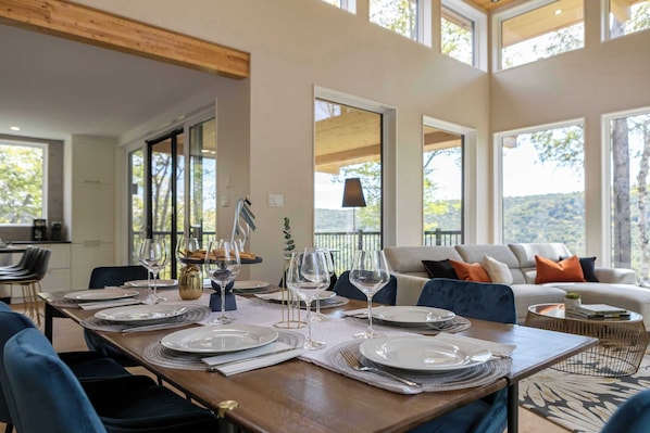 Un comedor con mesa de madera dispuesta para una comida, un sofá al fondo y ventanas amplias que ofrecen vistas al exterior.