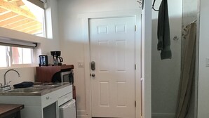 A compact kitchen with a sink, stove, and microwave. A white door leads to a bathroom with a shower.
