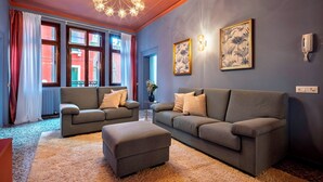 A modern living room with a grey sofa, a matching ottoman, and a chandelier. There are two framed botanical prints on the wall and a radiator near the window. - The Arsenal Large Apartment (Venice)