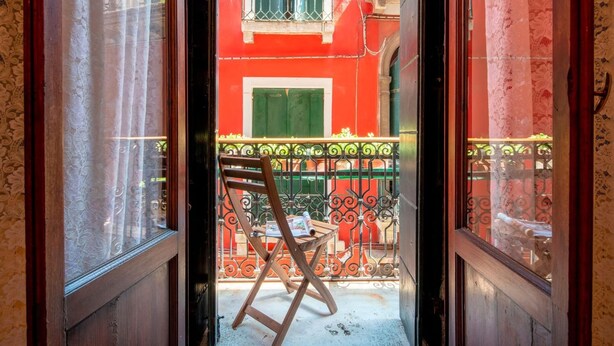 A view through a doorway into a courtyard with a wooden chair and a table, a red building with green shutters, and a metal railing. - The Arsenal Large Apartment (Venice)