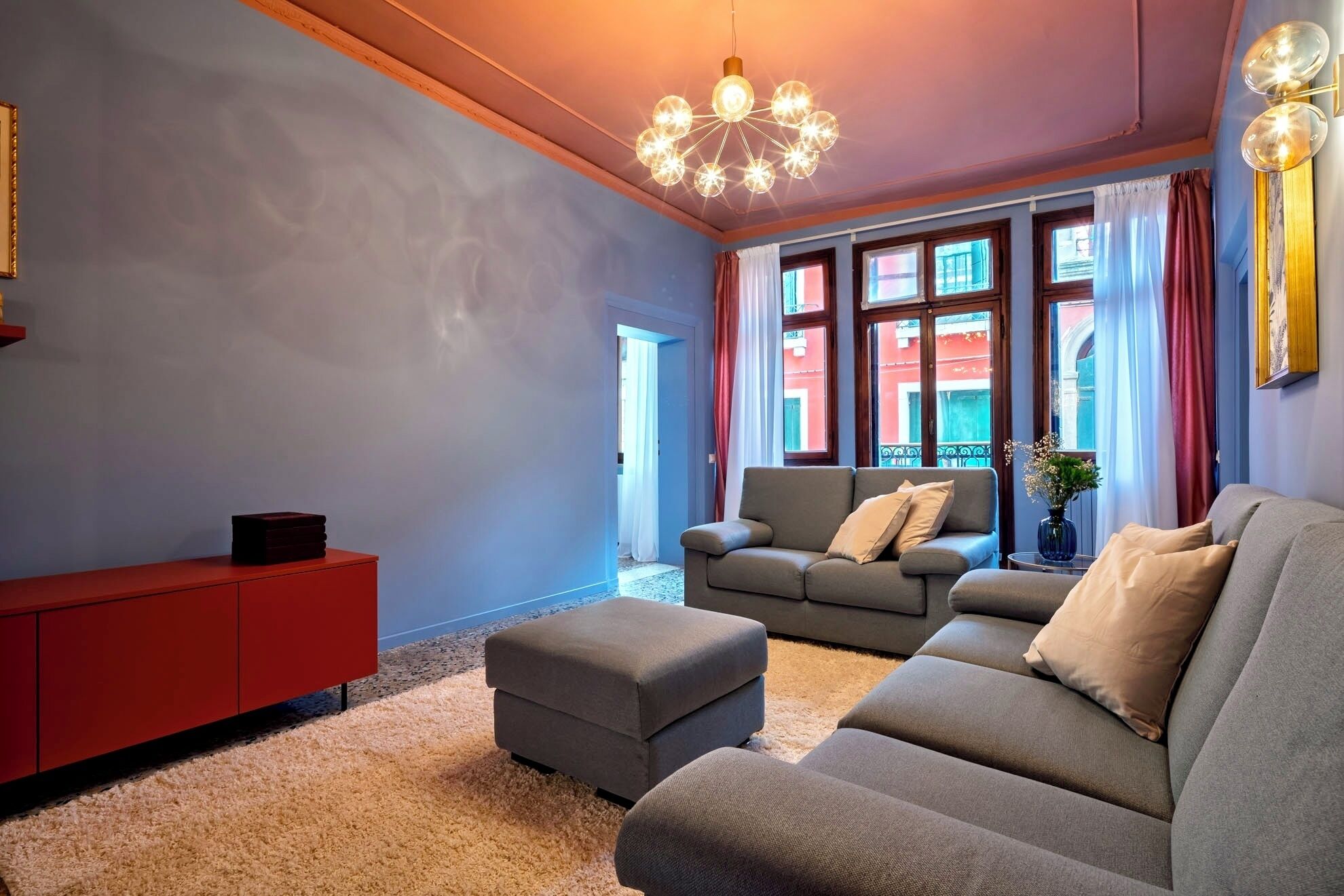 Ein Wohnzimmer mit einem roten Schrank, grauen Sofas, einer Kristallleuchter und großen Fenstern mit Vorhängen.