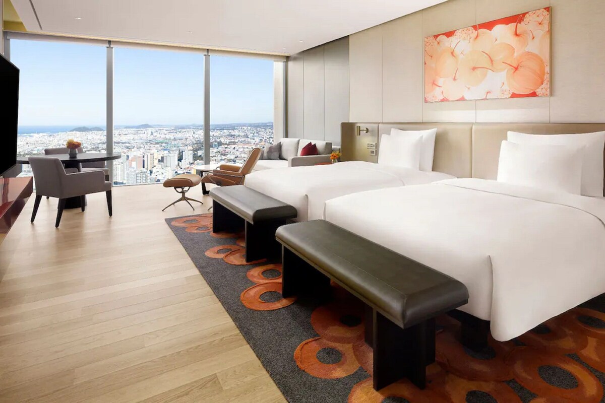 A hotel room with two beds, a desk, and a view of the city.
