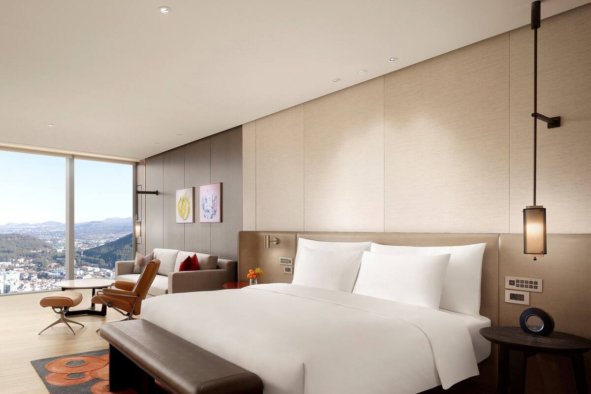 Ein modernes Hotelzimmer mit einem großen Bett, einer Couch, einer Bank und Blick auf die Stadt.