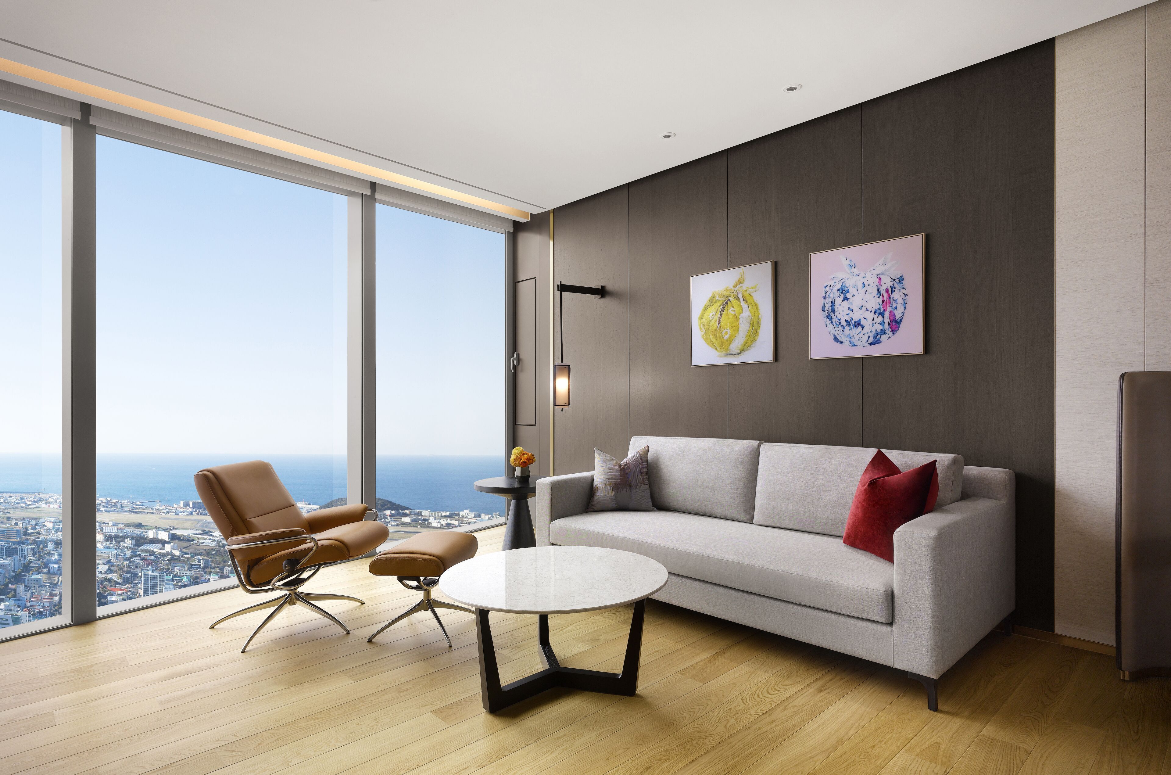 A modern living room with a grey sofa, brown armchair, and a round coffee table. There are two framed artworks on the wall, and a cityscape view through large windows.