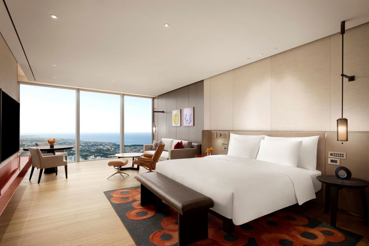 A modern hotel room with a large bed, a seating area with a table, and a view of the city.