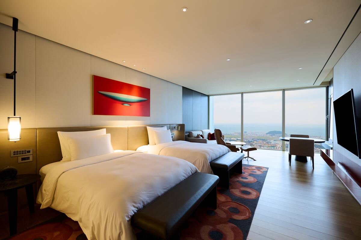 Ein modernes Hotelzimmer mit zwei Betten, einem großen Fenster mit Blick auf die Stadt, einem Flachbildfernseher und einem roten abstrakten Gemälde an der Wand.
