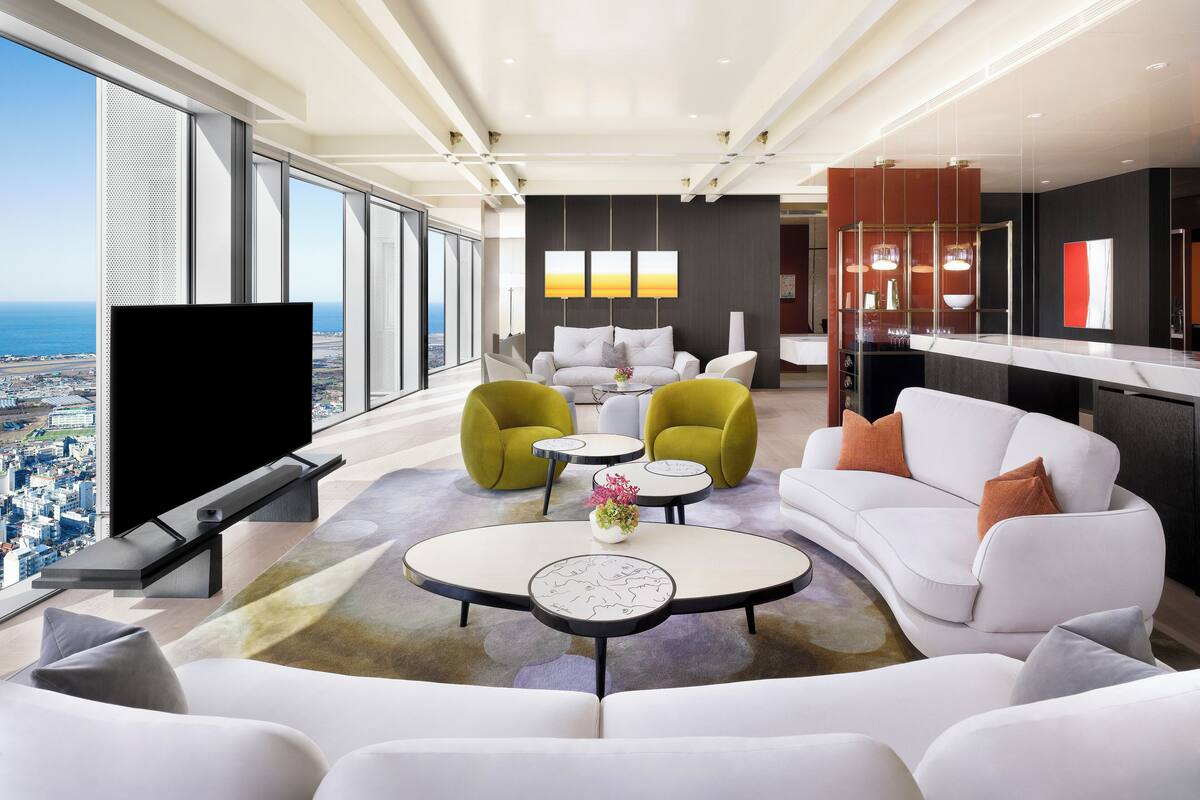 Ein modernes Wohnzimmer mit großem Fernseher, weißen Sofas, grünen Sesseln und einem Essbereich mit einem Glastisch.