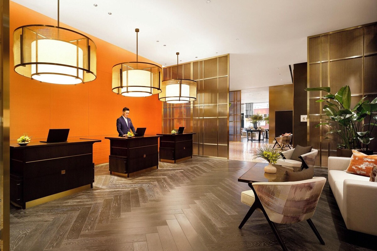 A hotel lobby with a reception desk, a seating area with a sofa and chairs, and large pendant lights.