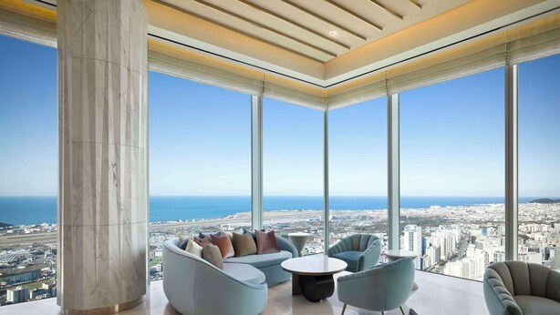 A high-rise interior with a panoramic ocean view, featuring a modern seating area with a sofa and armchairs, a coffee table, and a large column. - Grand Hyatt Jeju (Jeju City)