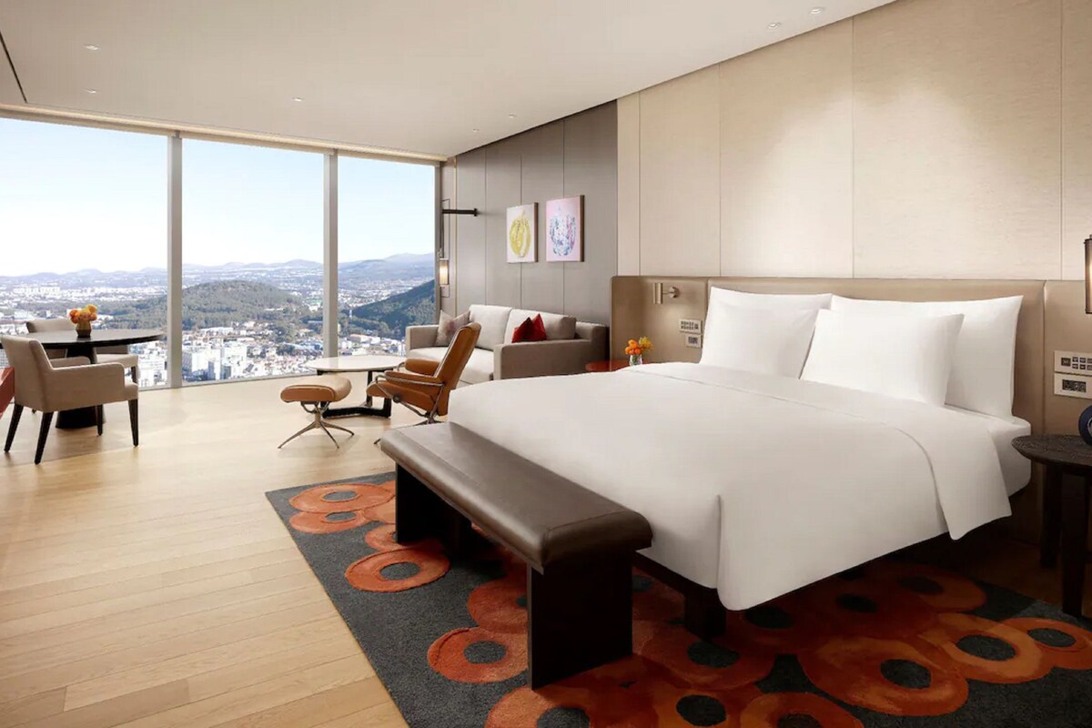 A modern hotel room with a large bed, a desk, and a chair. There is a view of a cityscape through large windows.