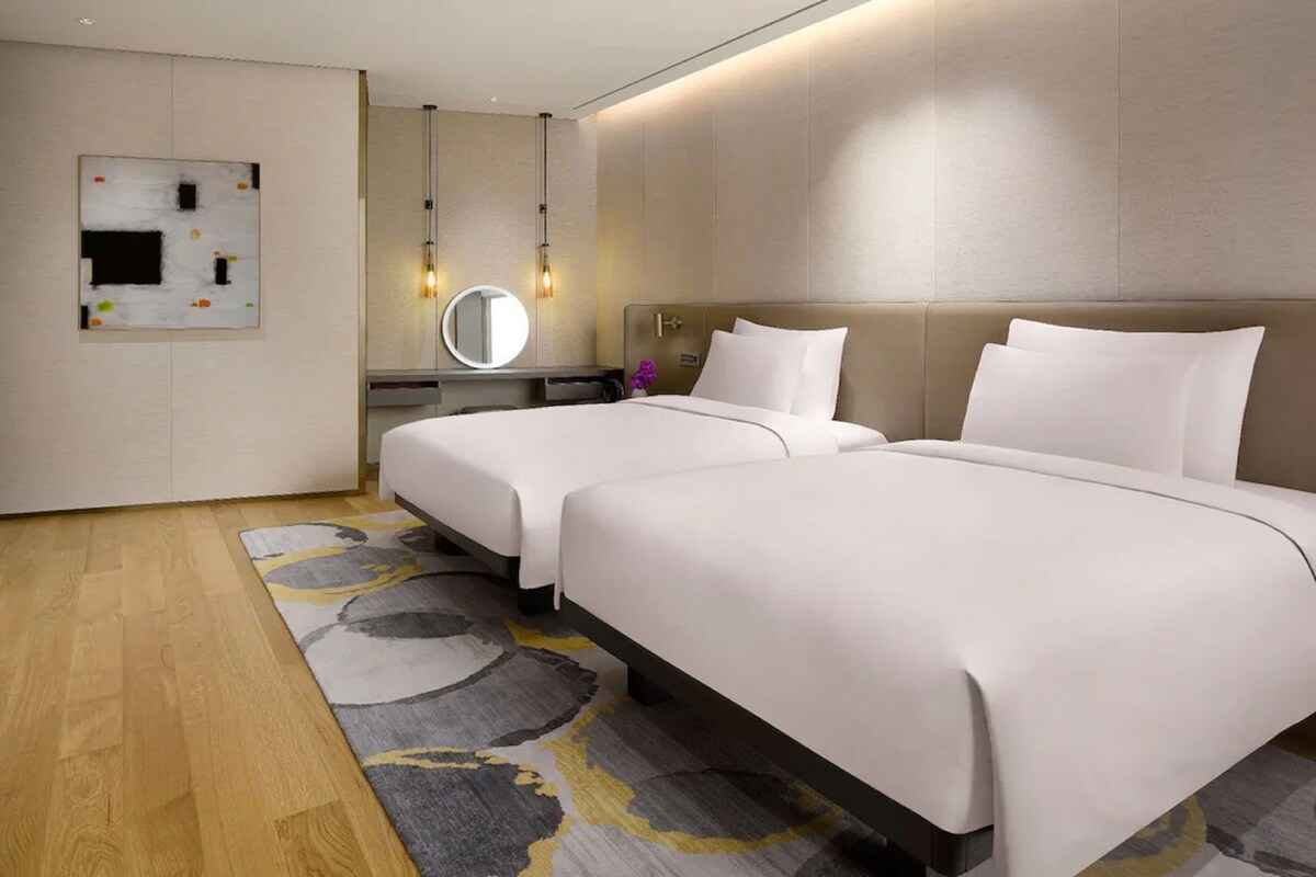 A hotel room with two beds, a TV, a desk with a mirror, and a modern light fixture.