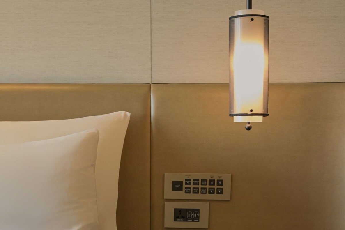 A hotel room with a bed, bedside table, lamp, and control panel.