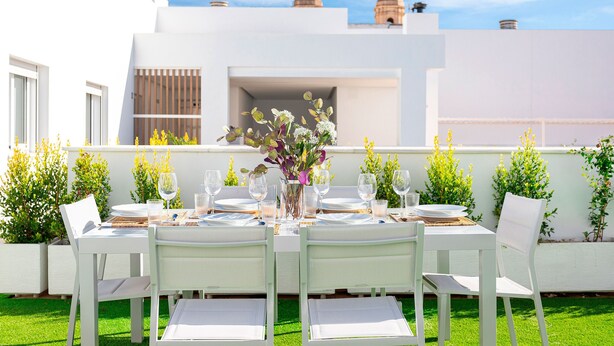 Un espace extérieur aménagé pour les repas, avec une table dressée pour quatre personnes, entourée de chaises blanches, et un vase rempli de fleurs placé au centre.