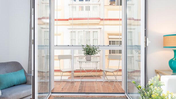 A balcony with a glass door, a grey sofa, a small table with a potted plant, and a view of neighboring buildings. - Genteel Home San Antonio Terrace II (Cadiz)