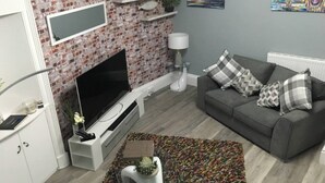 A living room with a brick wall, a flat-screen TV, a grey sofa with patterned cushions, a white side table, and a colorful rug. - Lovely 2- Bed Apartment in Hawick (Hawick)