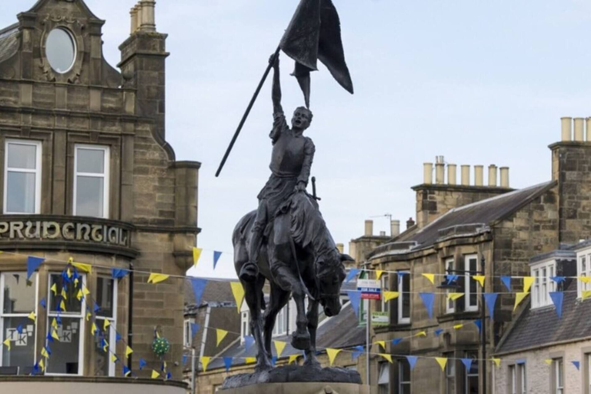 Eine Statue einer Person zu Pferd mit einer aufgerichteten Flagge, vor einer Kulisse aus historischen Gebäuden und blauem Himmel.