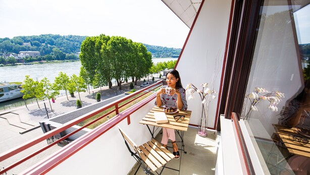 Un balcón con vista al río, una mesa con una silla y una persona usando un teléfono inteligente.