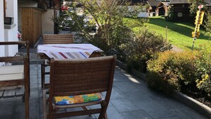 Et træbord og stole under en overdækket terrasse med udsigt til et bjerg og et kirktårn.