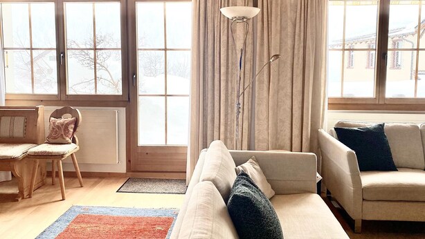 En hyggelig stue med en sofa, en lænestol og en gulvlampe. Store vinduer med gardiner, og udsigt til et sneklædt landskab udenfor.