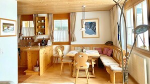 Et hyggeligt sommerhusinteriør med et træspisebord, stole, en bænk, et tekøkken og et stort vindue med gardiner.