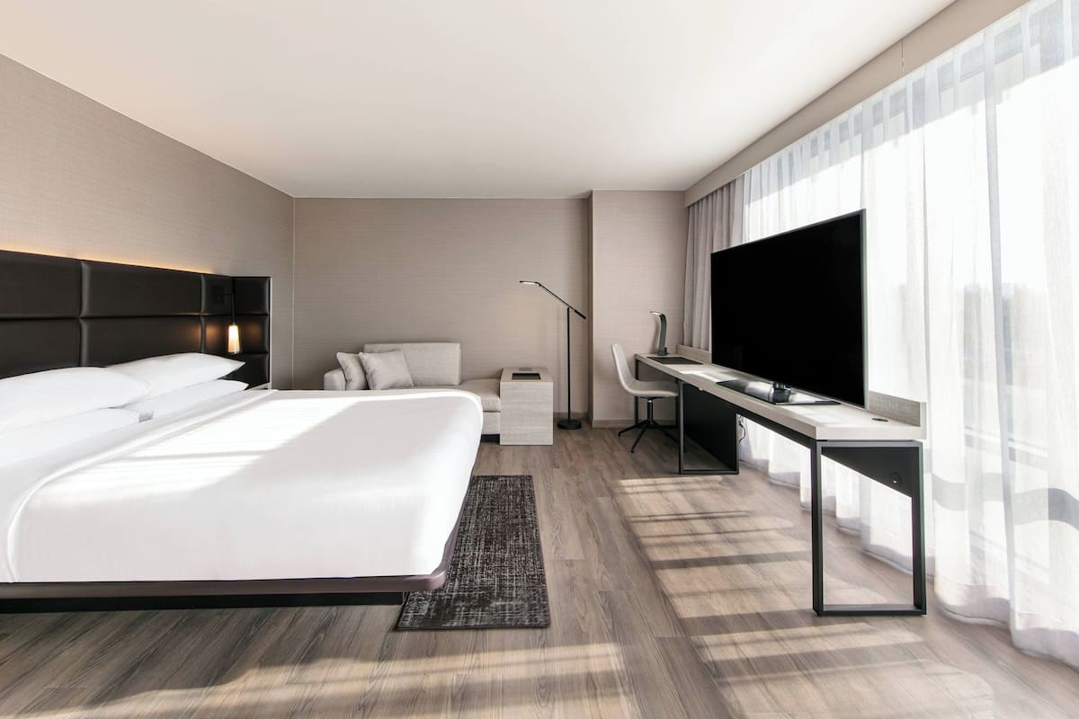 A modern hotel room with a large bed, a desk, a flat-screen TV, and a large window with sheer curtains.