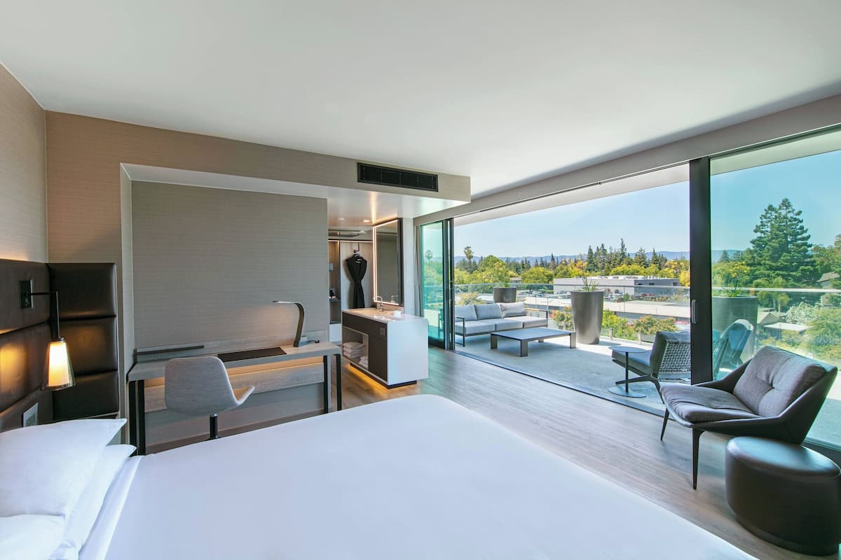 A modern hotel room with a large bed, a desk, a chair, and a balcony with a view of the city.
