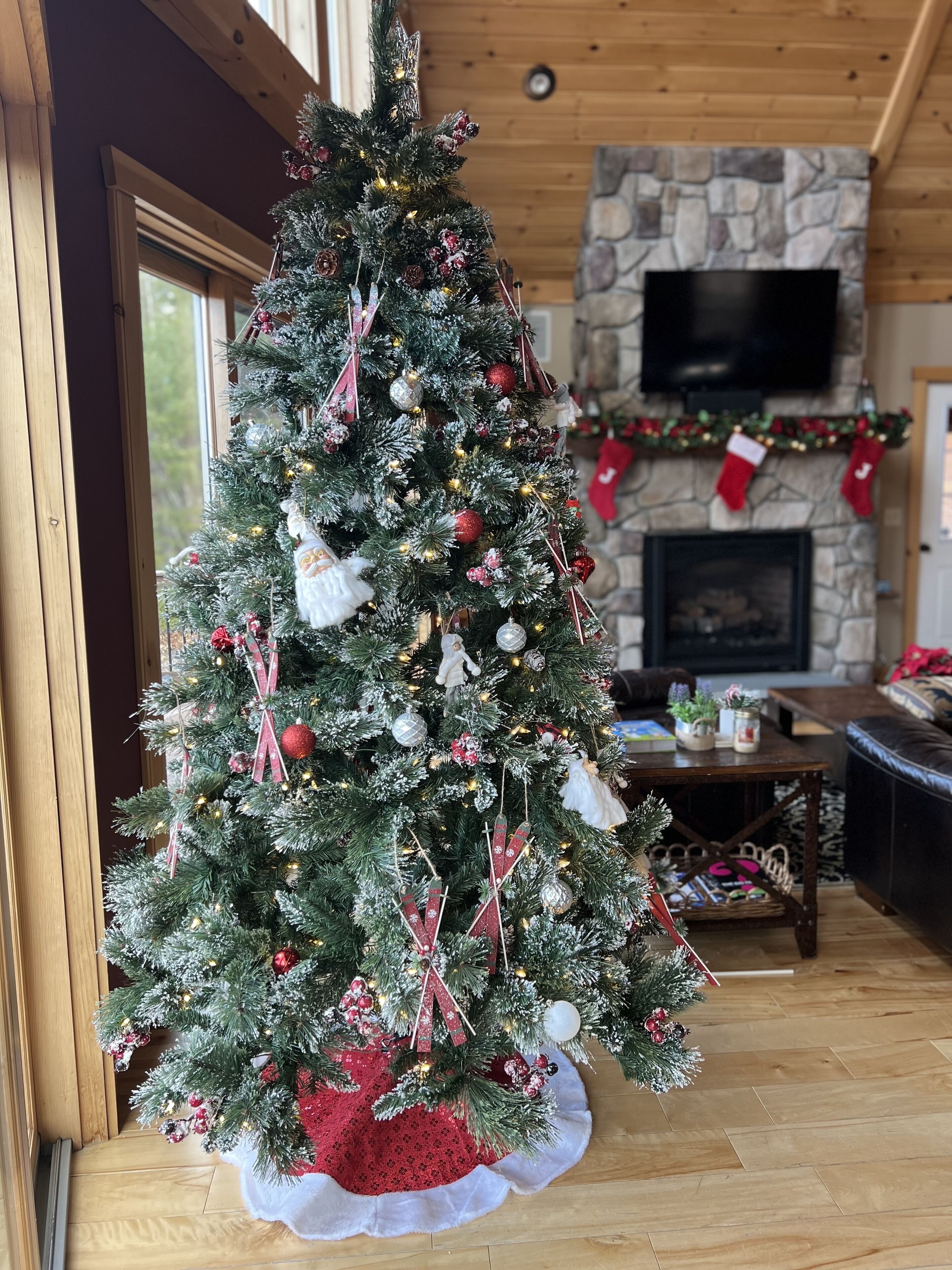 A Christmas tree with ornaments and lights, a stone fireplace, and a leather couch in a wooden interior.