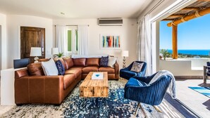 Ein modernes Wohnzimmer mit einer braunen Ledersofa, einem Holztisch und einem blauen Sessel. Ein großes Fenster bietet Blick auf den Ozean.