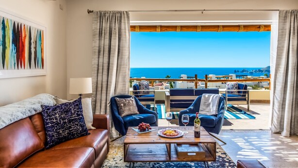 Ein Wohnzimmer mit Meerblick, einem Holztisch mit Früchten und Wein sowie blauen Polsterstühlen.
