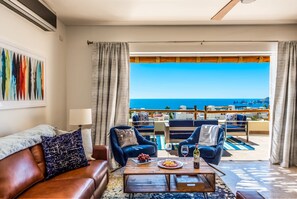 Sala de estar com vista para o mar, mesa de centro de madeira com frutas e vinho, e poltronas estofadas em azul.