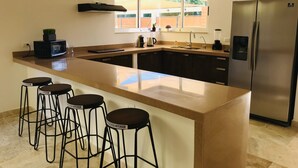 Een moderne keuken met een centraal eiland, barstoelen, een koelkast en een raam met uitzicht op een achtertuin.