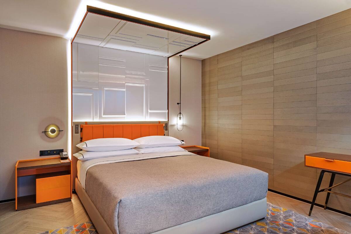 Una habitación de hotel moderna con una cama grande, una mesita de noche, un escritorio y un espejo fijado a la pared.