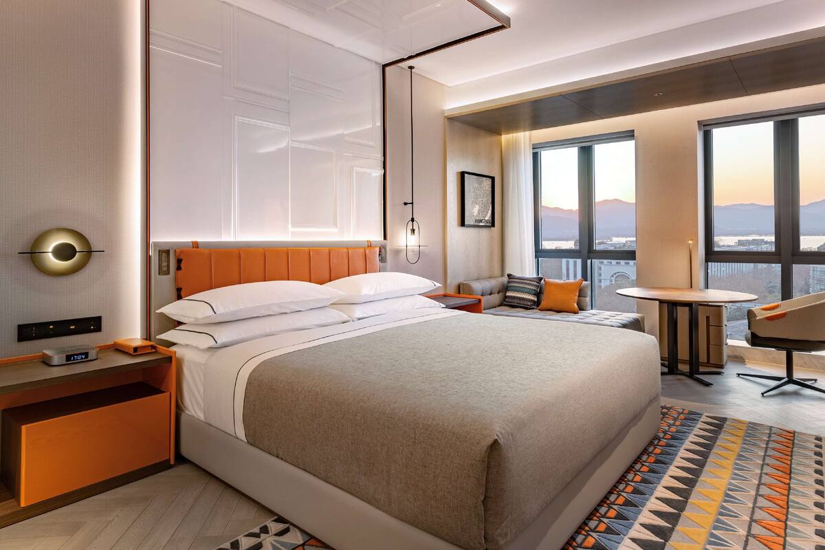 Una habitación de hotel moderna con una cama grande, una mesita de noche con lámpara, un pequeño rincón de descanso y vistas a la ciudad a través de amplios ventanales.
