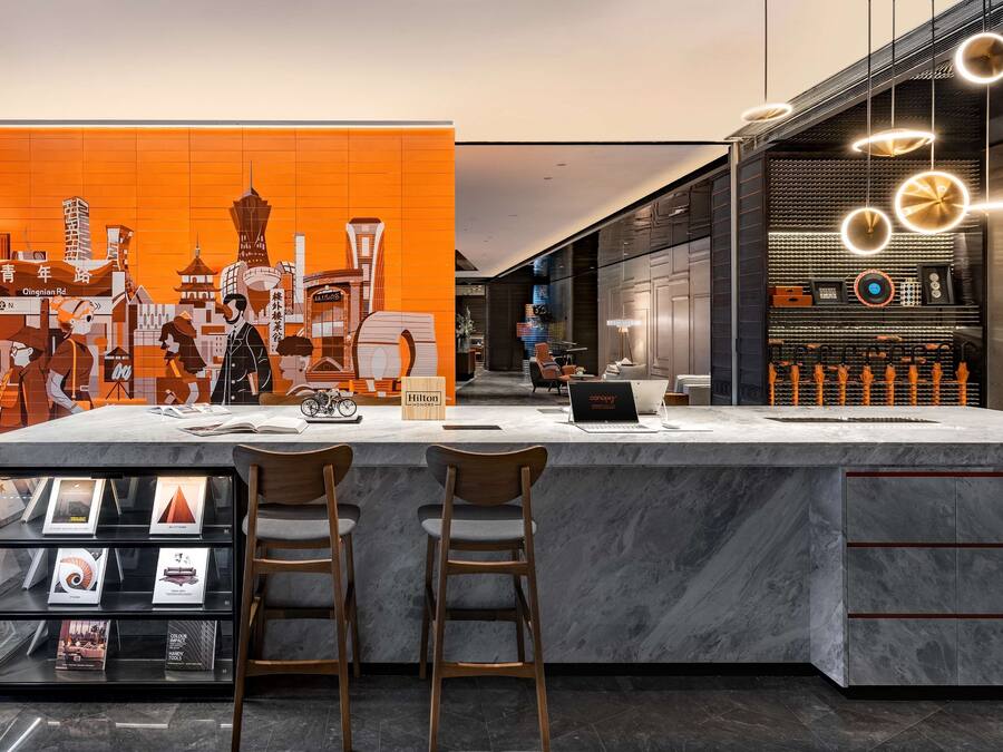 Un moderno vestíbulo de hotel con una recepción de mármol, dos taburetes de bar, una pantalla digital y un mural con vistas a la ciudad.