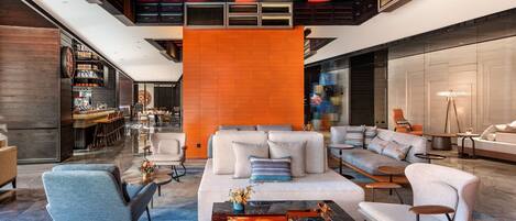 A spacious lobby with a modern design, featuring a large seating area with sofas and armchairs, a bar area with shelves, and a unique geometric ceiling design.