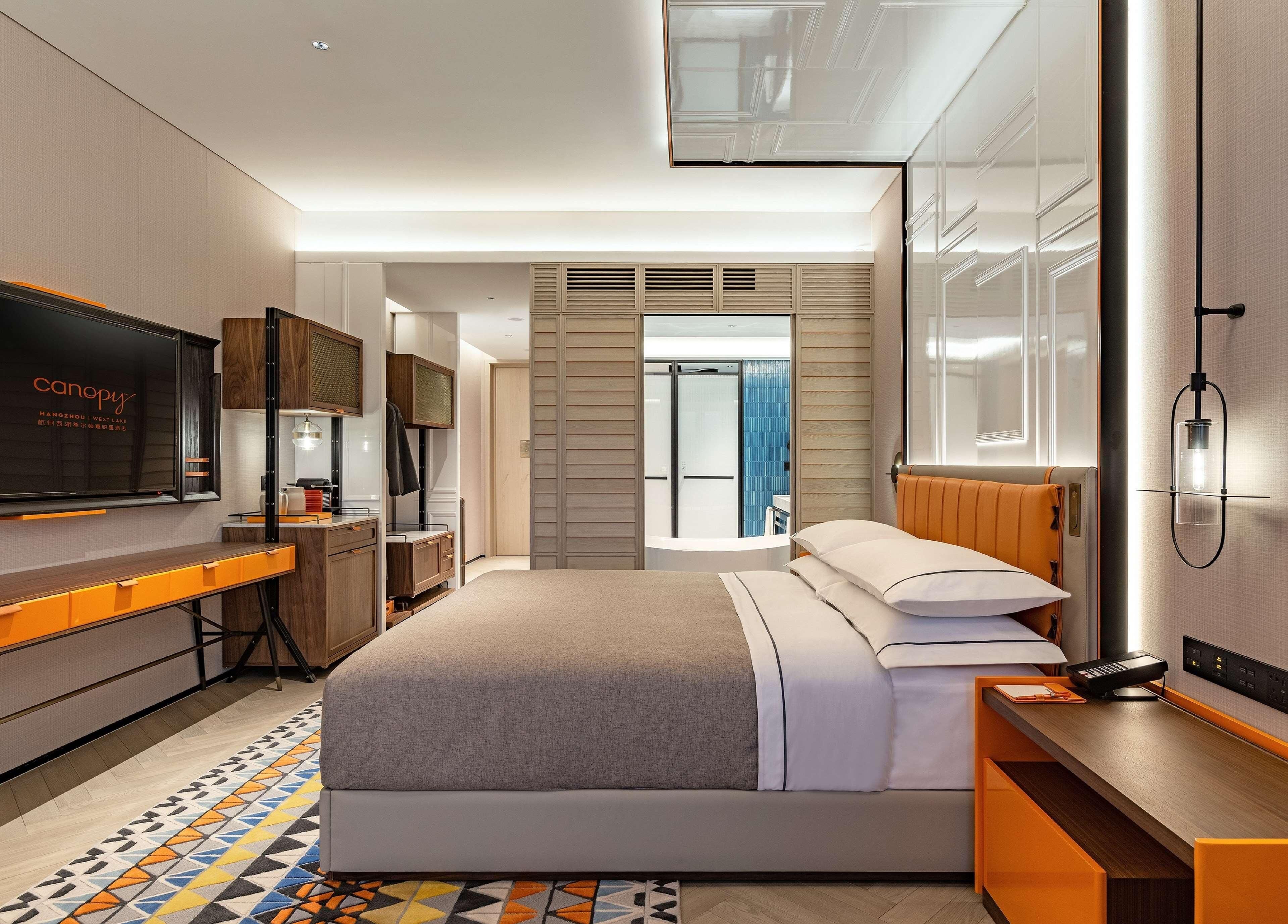 A modern hotel room with a bed, a desk, a TV, and a bathroom visible through a glass door.
