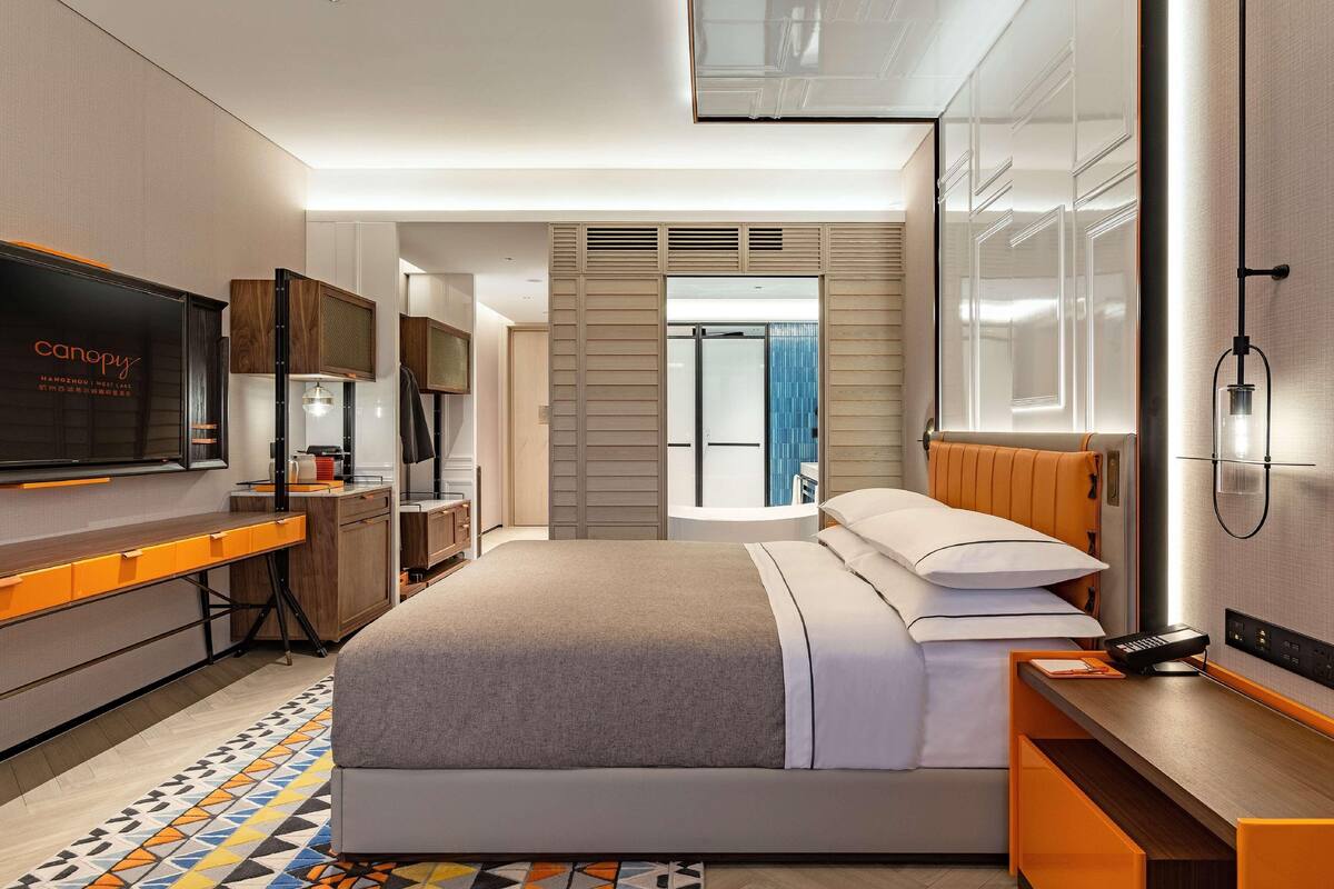 Habitación de hotel moderna con cama, escritorio, televisor y baño visible a través de una puerta de cristal.