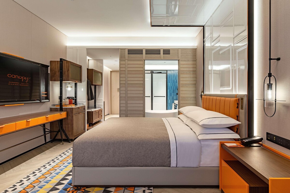 Quarto de hotel moderno com uma cama, uma escrivaninha, uma televisão e um banheiro visível por uma porta de vidro.