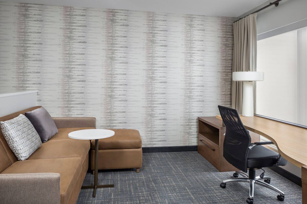 A hotel room with a sofa, a small round table, a desk with a chair, a lamp, and a large window.