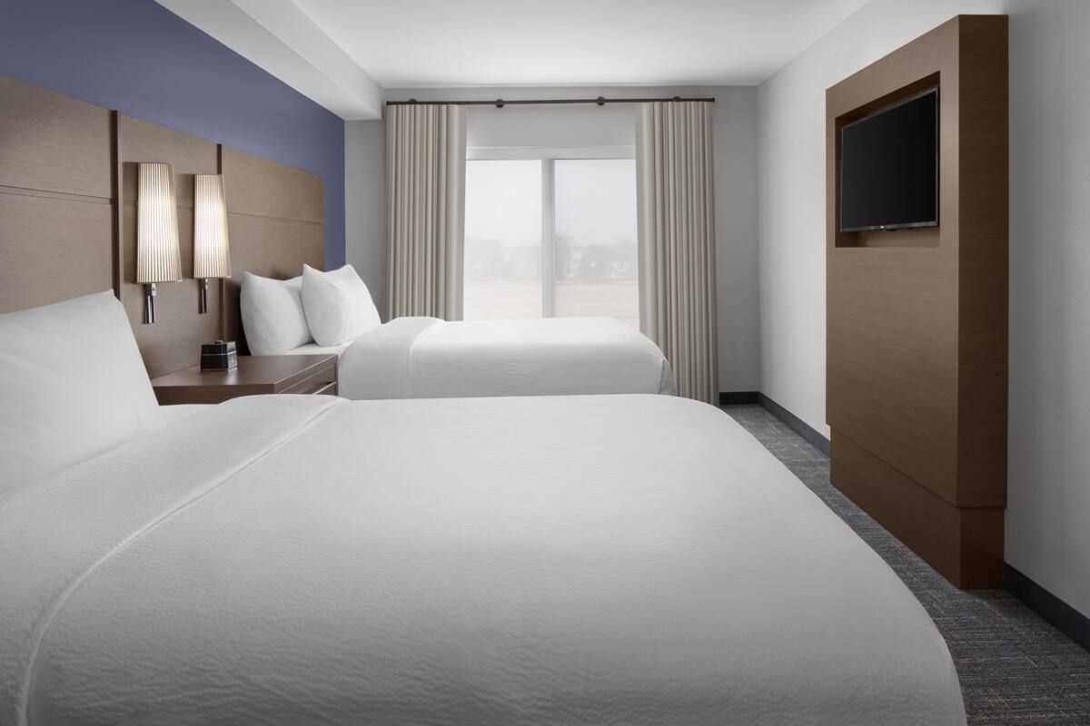 A hotel room with two beds, a TV mounted on the wall, and a large window with curtains.