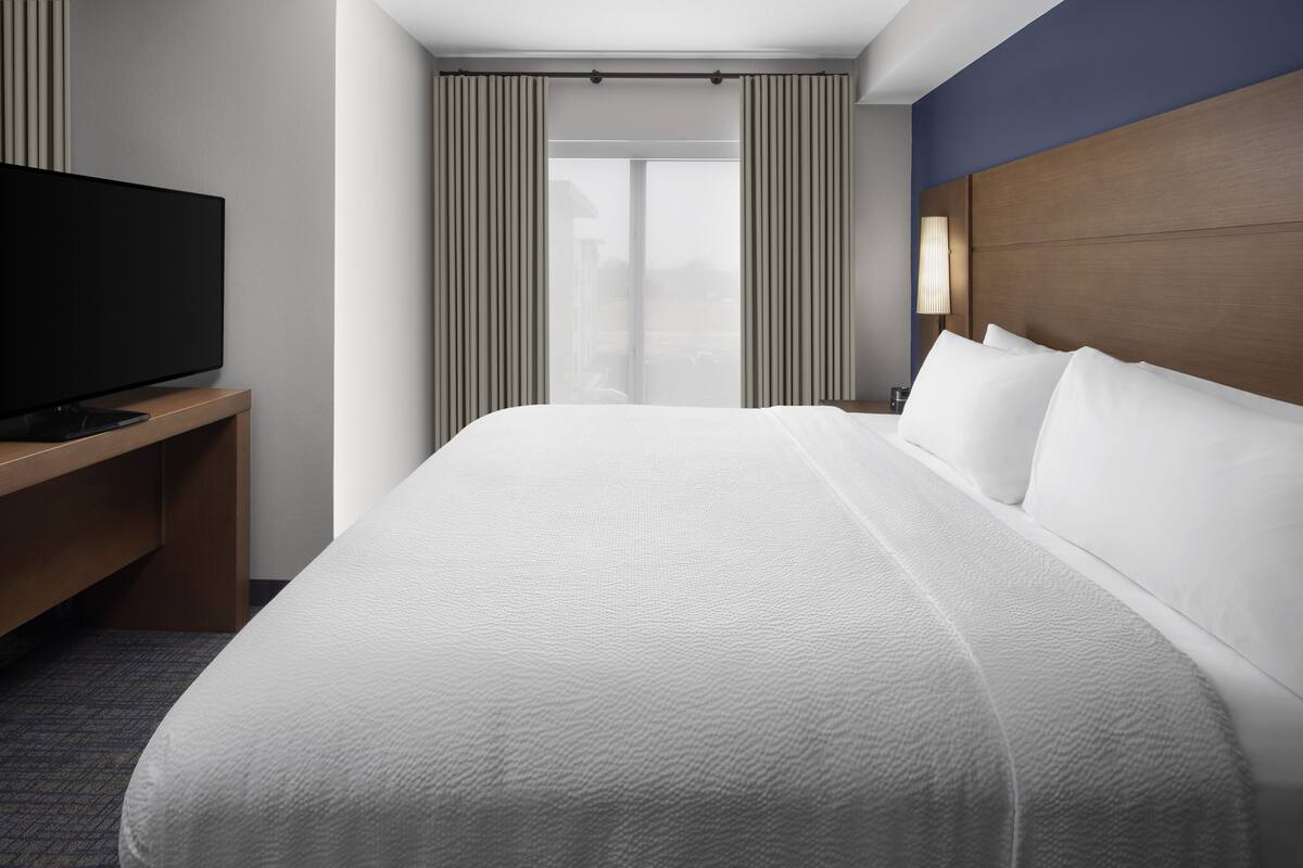 A hotel room with a large bed, a flat-screen TV on a wooden stand, a nightstand with a lamp, and a view of the outdoors through a large window with curtains.