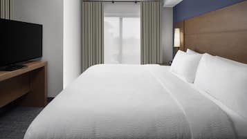 Habitación de hotel con una cama grande, un televisor de pantalla plana sobre un soporte de madera, una mesita de noche con lámpara y vistas al exterior a través de un ventanal con cortinas.