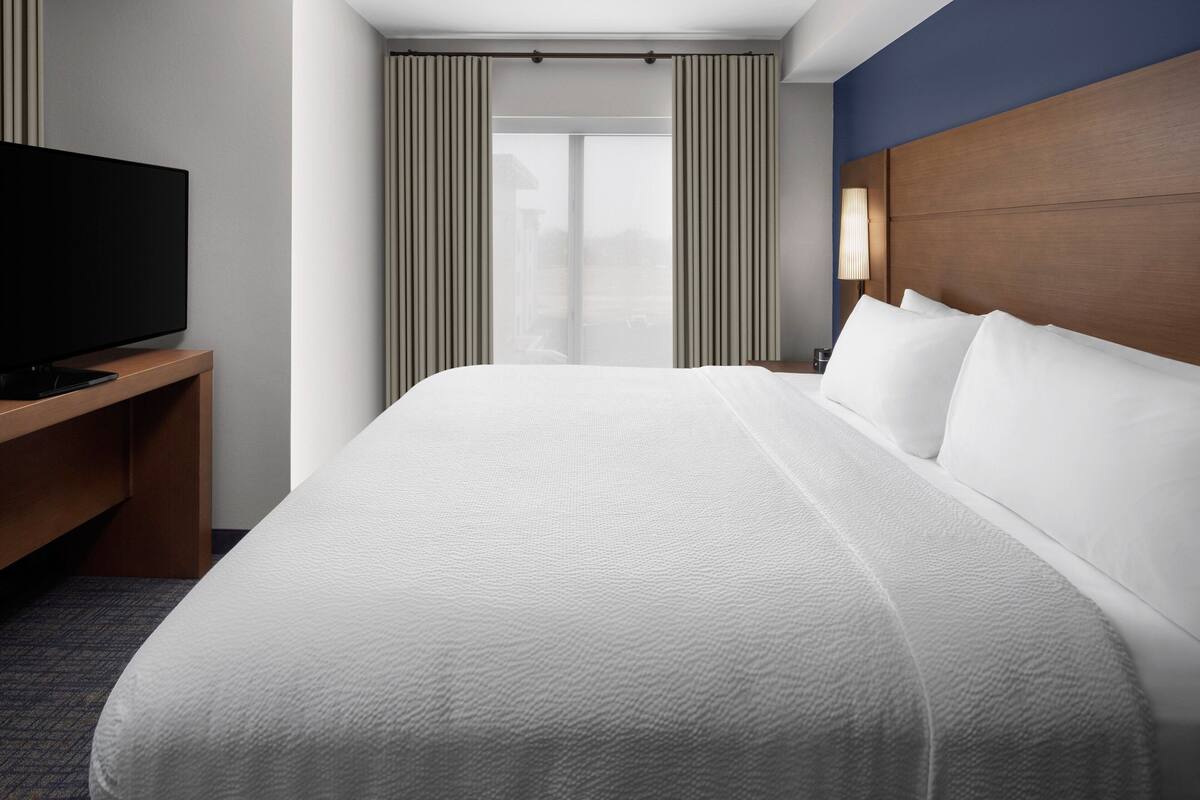 A hotel room with a large bed, a TV on a wooden stand, and a view of the outside through a window with curtains.