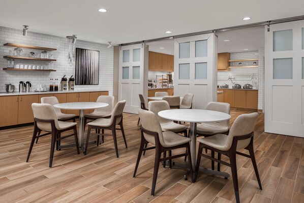 Une cuisine moderne avec des meubles blancs, des plans de travail en bois et un îlot central. On y trouve plusieurs tables rondes avec des chaises, et une porte coulissante donne accès à un coin repas.