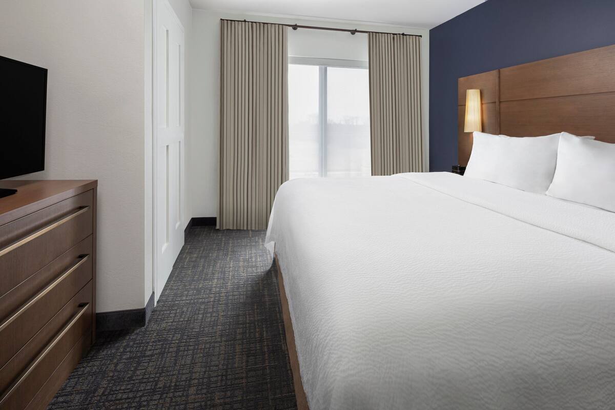A hotel room with a large bed, a TV on a wooden dresser, a window with curtains, and a nightstand with a lamp.