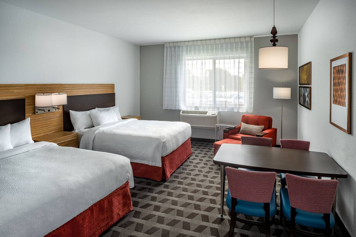 A hotel room with two beds, a dining table, chairs, a sofa, a window with curtains, and a hanging light fixture.