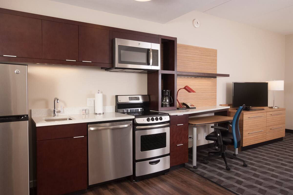 A compact kitchen with a microwave, stove, and refrigerator. There is a small dining area with a table and chair, and a TV on the wall.